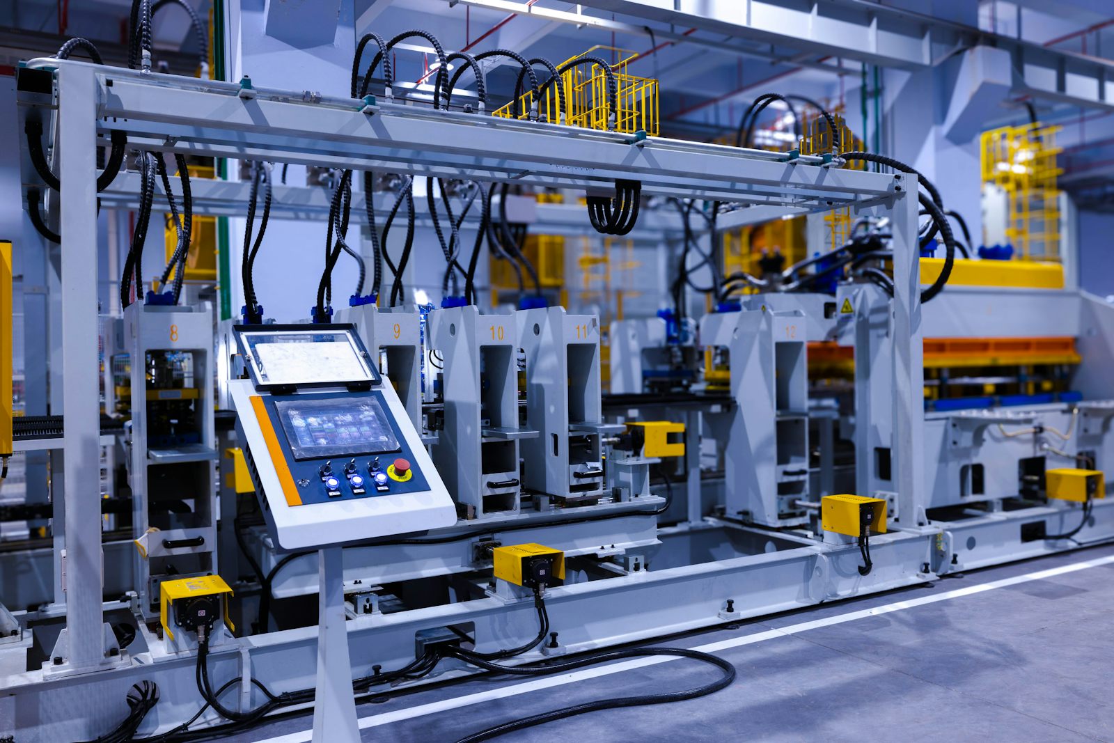 An operator touchscreen and control buttons on an automated forming line in a factory, with numbered stations and sensor cables behind it