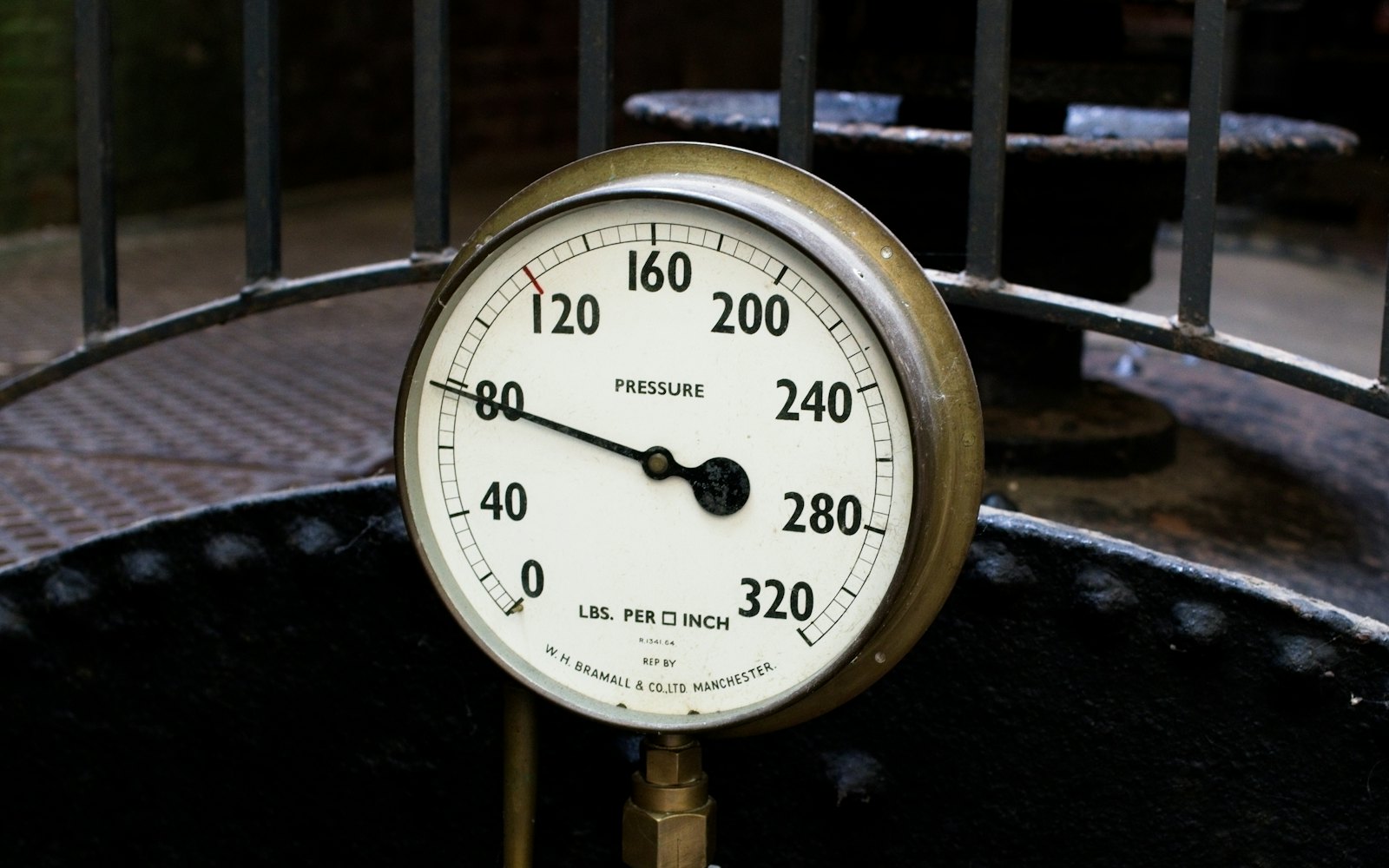 A brass pressure gauge reading about 80 pounds per square inch on old black machinery at a textile mill