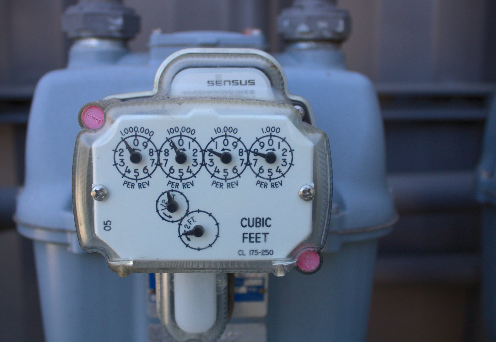 A grey utility gas meter with four round dials counting cubic feet, mounted outdoors on a pipe