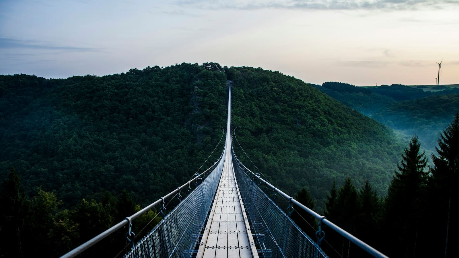 Long empty suspension footbridge stretching toward a dark green forested hill at dusk