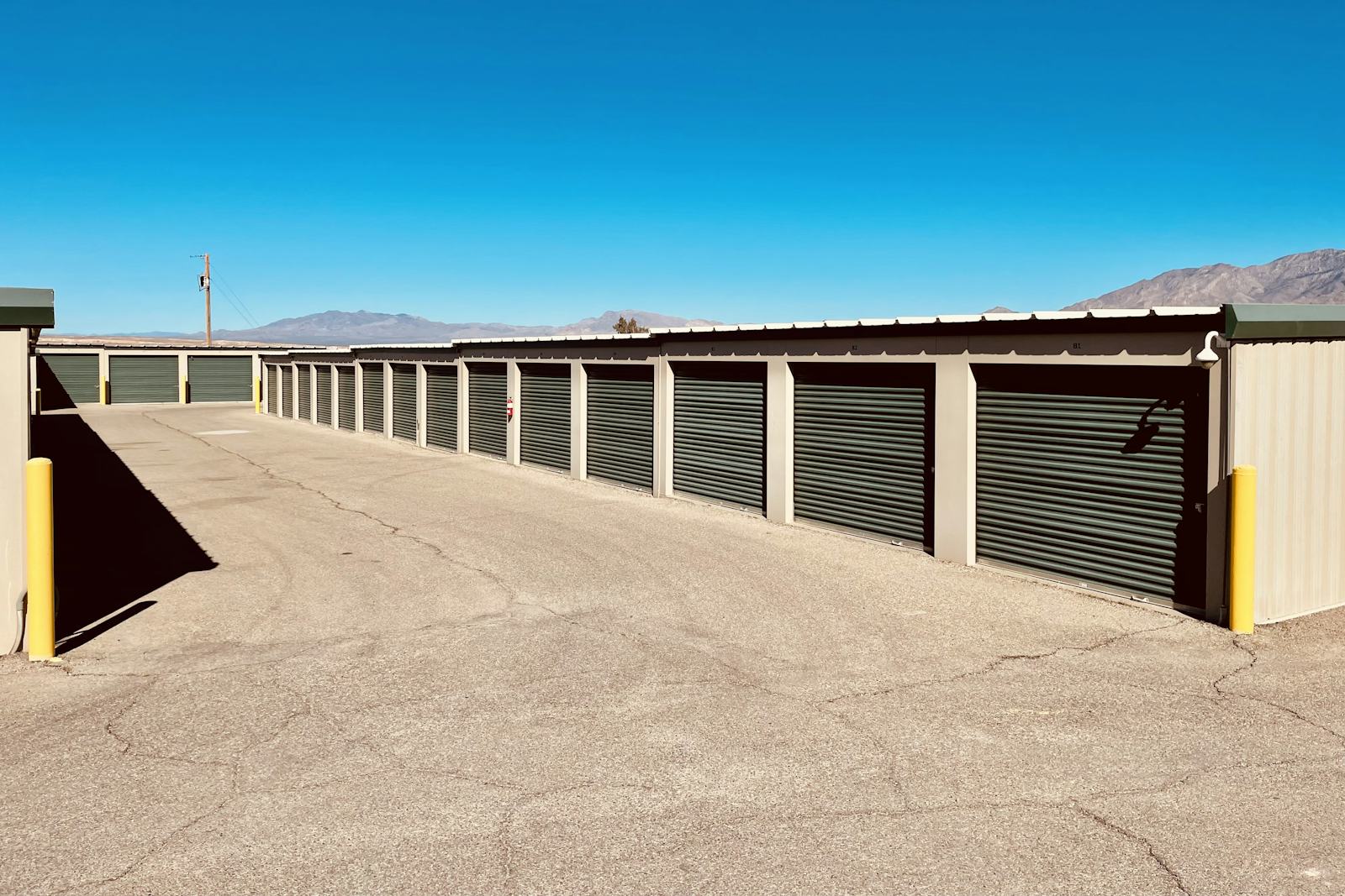 A long row of identical self-storage units with closed green roll-up doors under a clear blue sky, desert mountains behind