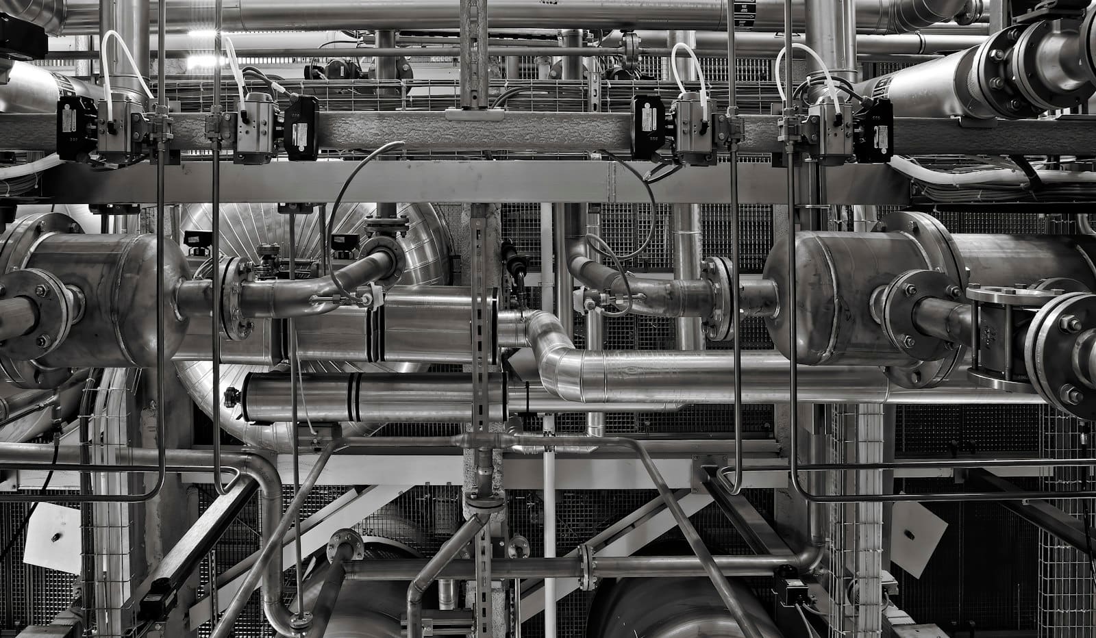 A dense network of grey industrial metal pipes and valves running in every direction through a factory