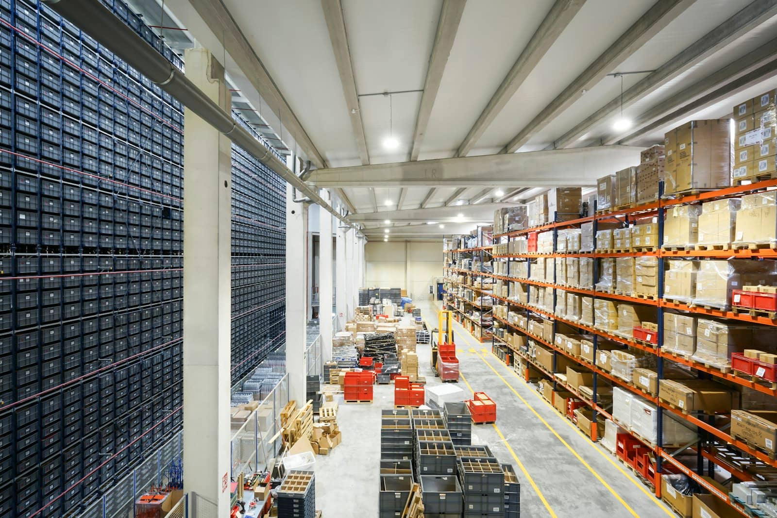 A large storage warehouse with every ceiling light on and no one in it, orange pallet racks of boxes on one side and a wall of grey crates on the other