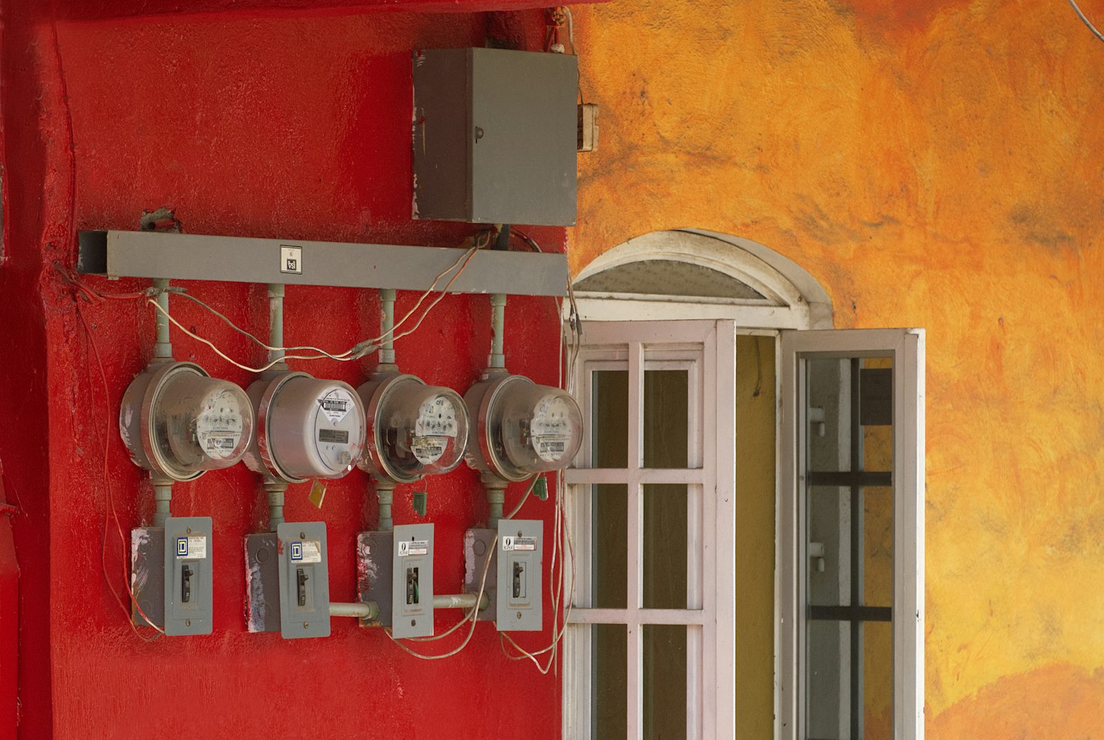 Four round electricity meters with their switch boxes mounted in a row on a red wall, wired to one supply bar beside an open window