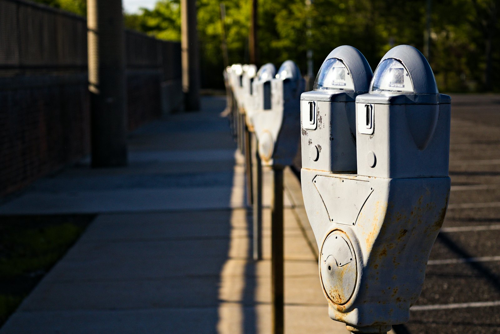 A row of grey coin parking meters along a sidewalk in late sun, beside an empty parking lot with painted bays