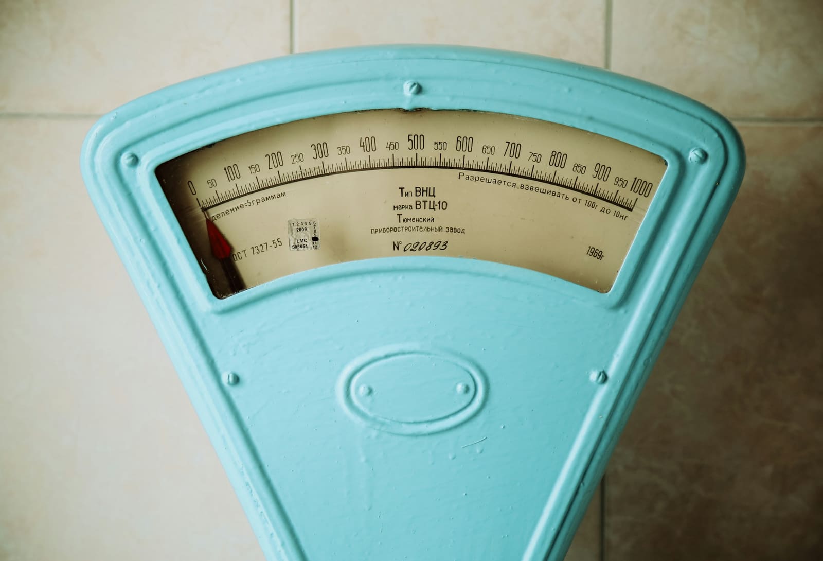A vintage pale blue weighing scale with a dial marked from 0 to 1000 grams, its needle resting at zero, against a tiled wall