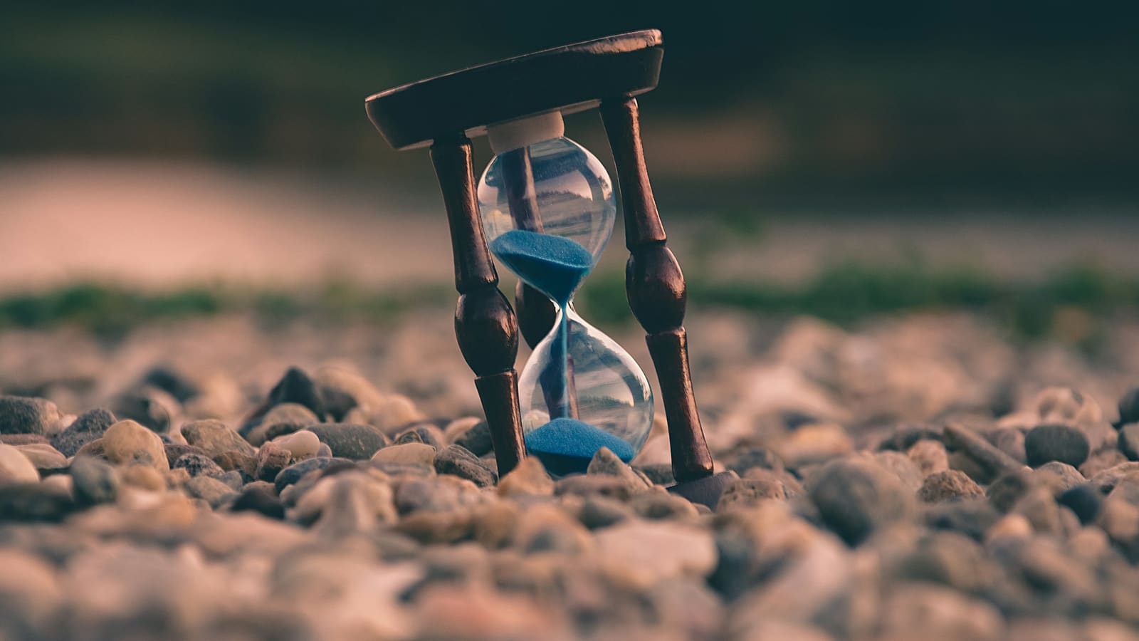 Wooden hourglass with blue sand standing among pebbles with a blurred background