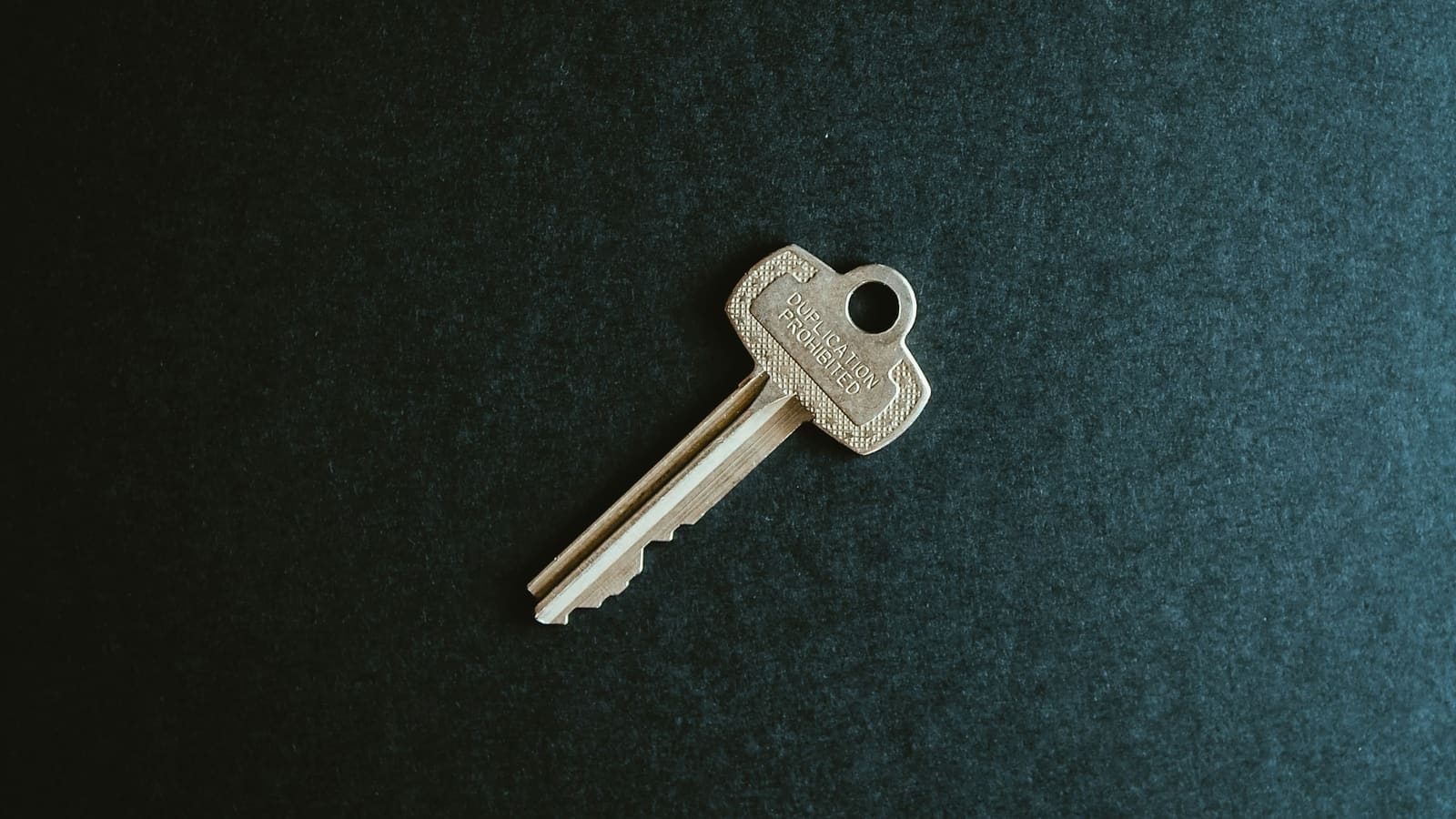 Single brass key lying on dark teal textured paper seen from above