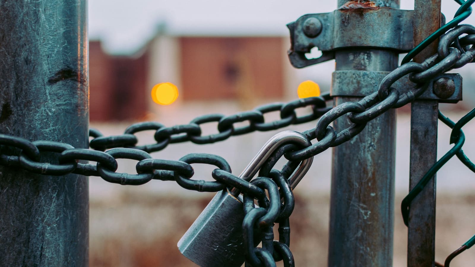 Steel padlock and heavy chain securing a metal gate post, blurred lights behind