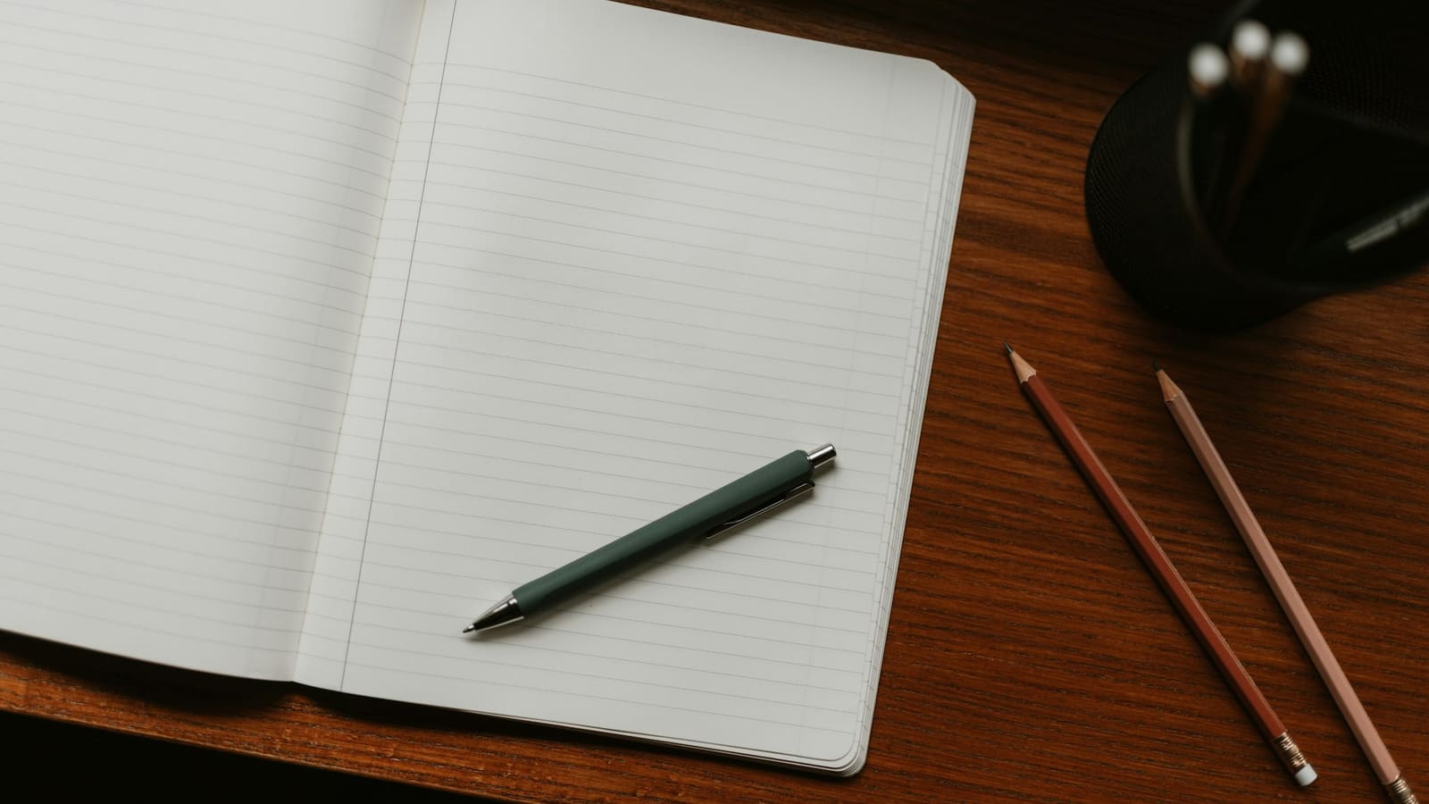 Open blank lined notebook with a green pen and pencils on a wooden desk